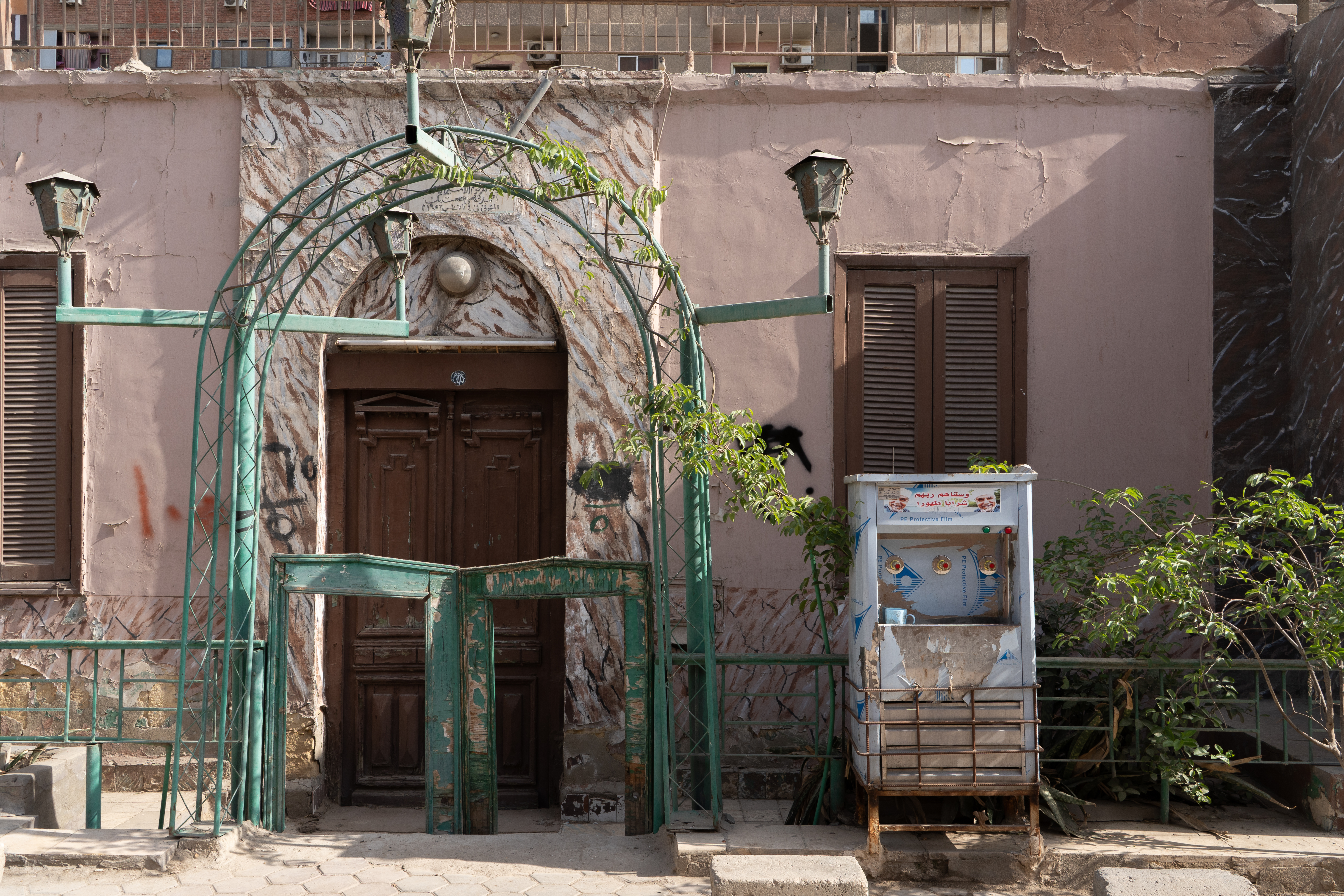 A kuldīr with three taps sits on a street outside of a pink building next to a green metal archway.