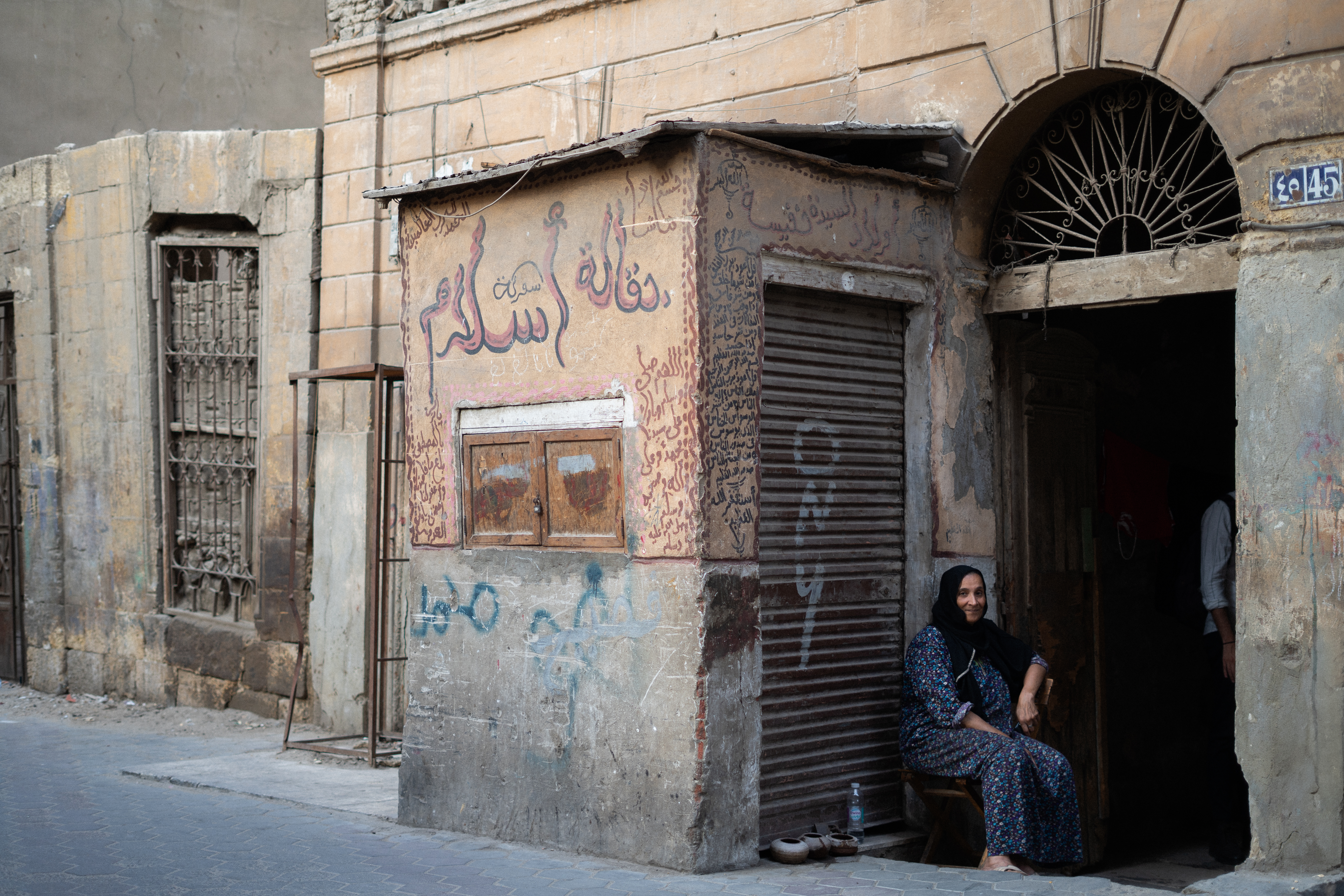 A slightly smiling woman wearing a black hijab looks at the camera and sits outside of a doorway with sabīls for birds resting on the ground in Khalifa.
