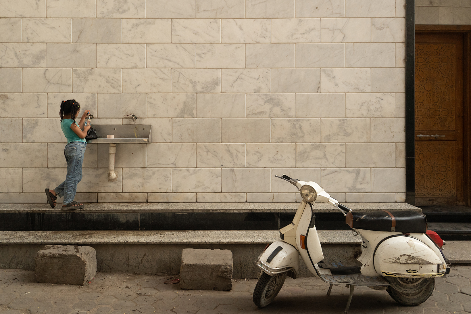 A young girl fills a water bottle from a faucet mounted on an exterior white marble wall in the Khalifa neighborhood. A white scooter sits in the foreground to the right.