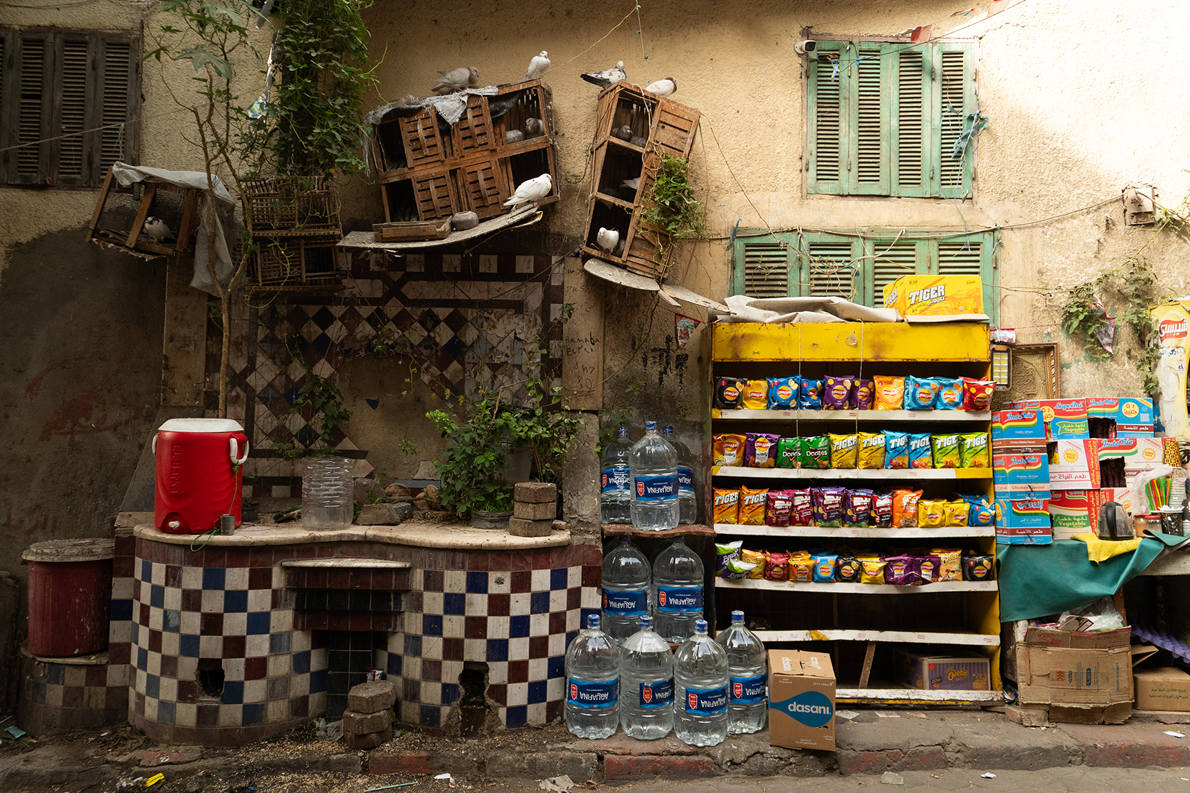 A display of snacks and water on a street in Khalifa. There is a red kūlmān for people as well as a dovecote and sabīl for birds.